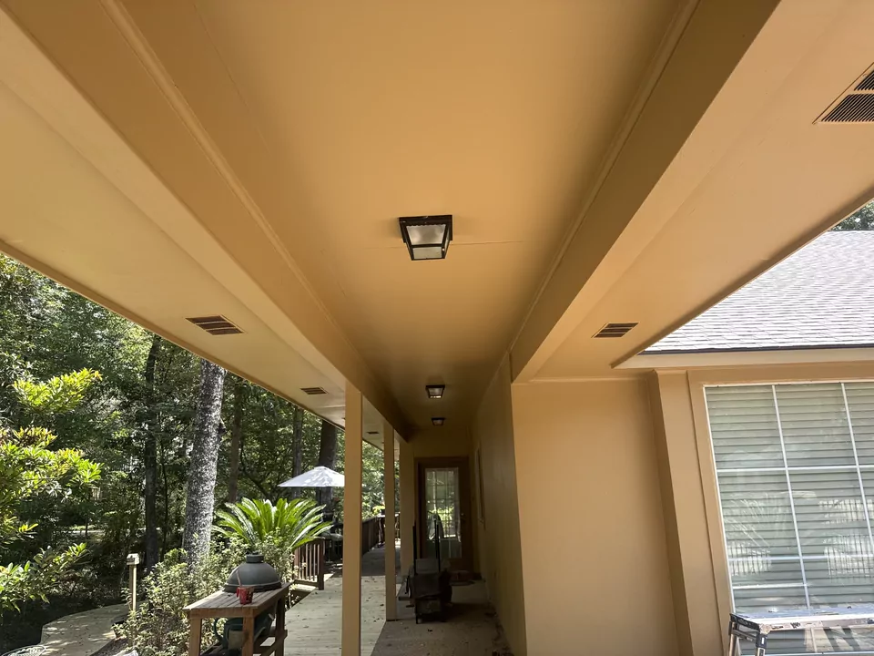 A long covered porch seen down its length, the ceiling and beam freshly painted, with lantern fixtures and woodland beyond the rail.