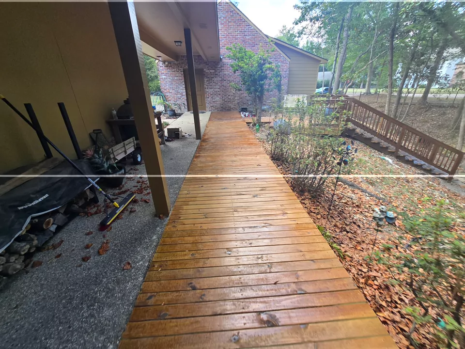 A long wooden walkway running alongside a brick house, freshly stained a warm golden tone, with autumn leaves scattered across it.