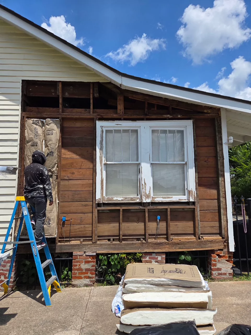 An exterior wall opened up for repair: siding removed around a window to expose the framing and sheathing, with a worker on a ladder at the left.